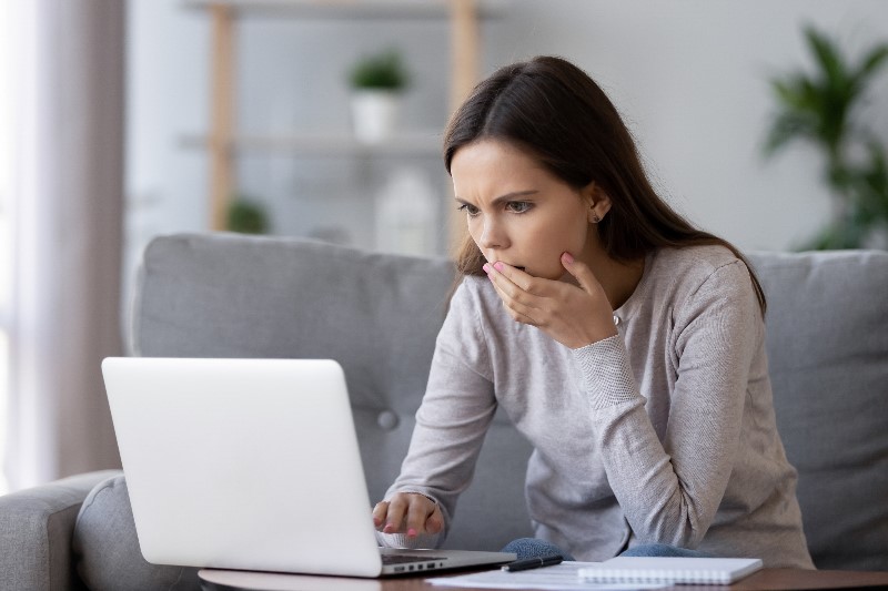 Young woman looking at her laptop with her hand over her mouth and a concerned expression on her face.